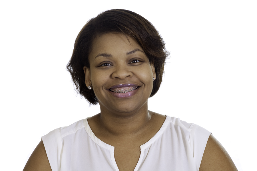 This Brian Charles Steel photo is a headshot of a woman in a white top.  She is of color, and she is centered in the frame.  Her hair is straight and combed to the right. She is smiling and has braces on her teeth.  She is lit in a slight Rembrandt style with one side slightly lighter than the other.  The background is completely white. 